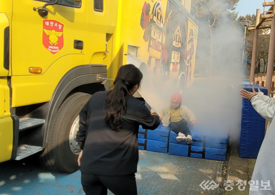 ▲ 이동형 체험차량에서 뿜어져 나온 연기 속으로 어린이가 대피하는 장면을 보호자가 지켜보고 있으며, 현장 참여자들이 실전과 유사한 상황에서 탈출 요령을 익히고 있다